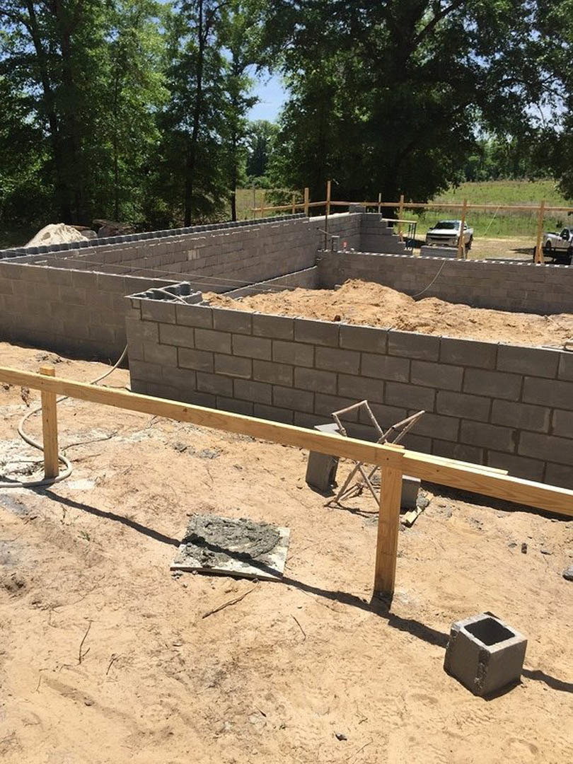 Concrete foundation slab surrounded by dirt and construction materials, wooden framing, metal fence, and white truck parked beside the site.