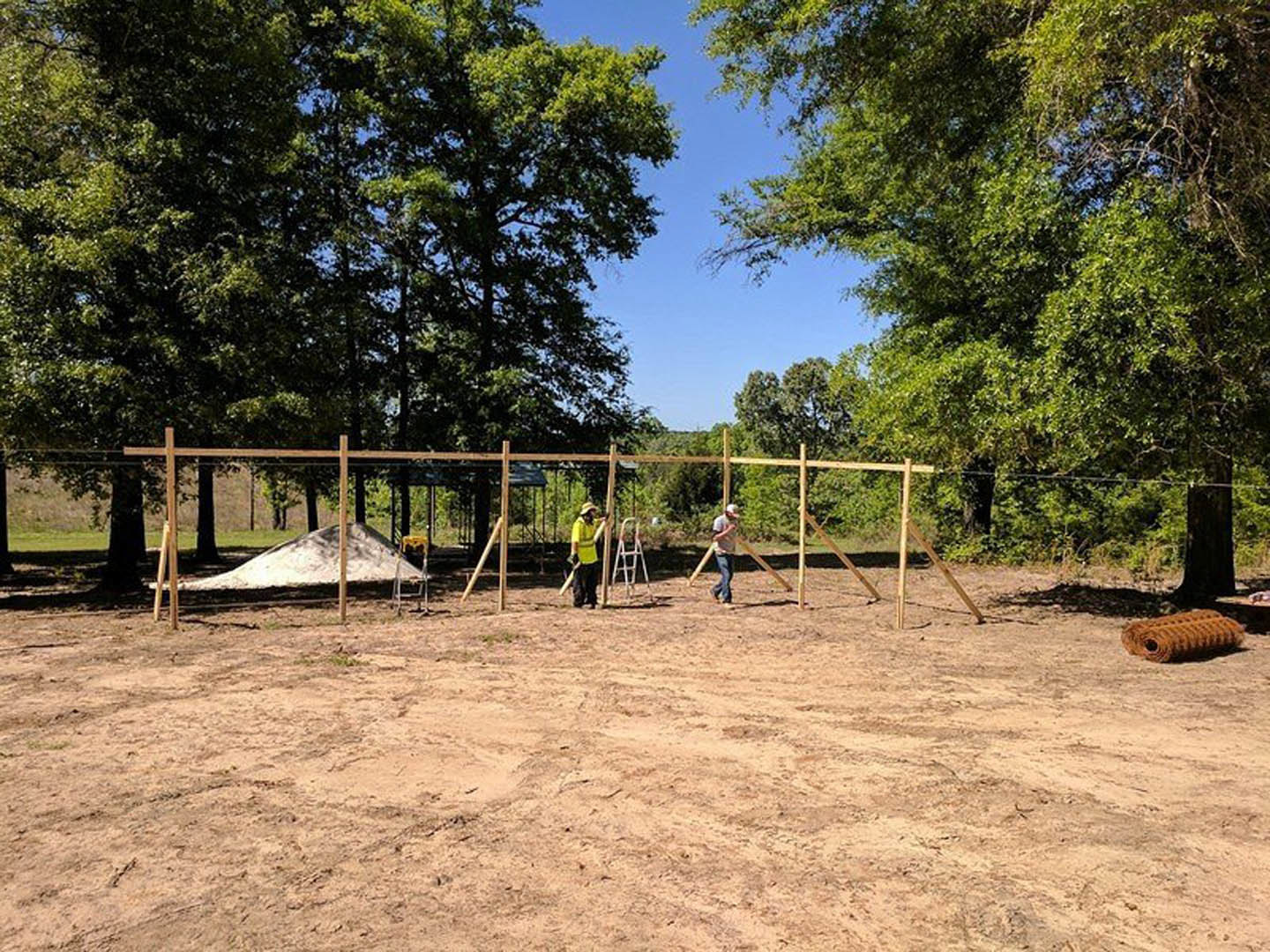 Crew building custom home foundation with exposed dirt, construction materials, and surrounding trees under clear sky