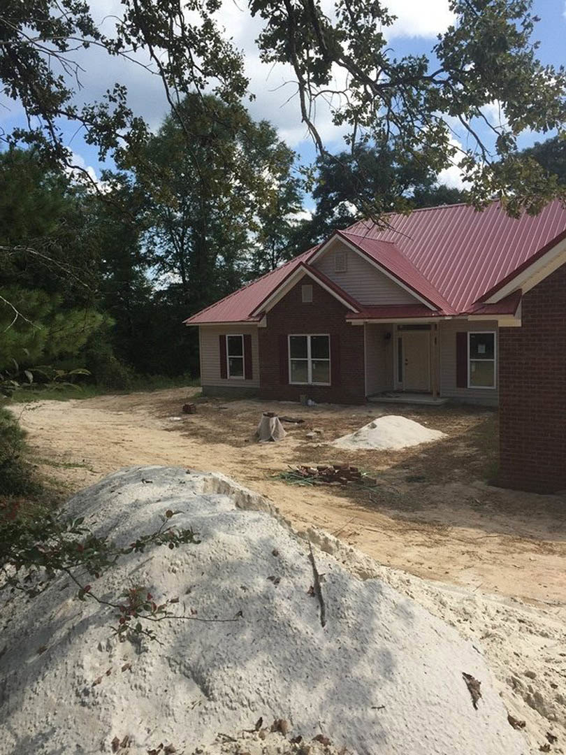 Red metal roof atop a white cottage-style home, white-framed window visible, surrounded by sandy ground and scattered branches, tree in background under clear sky.