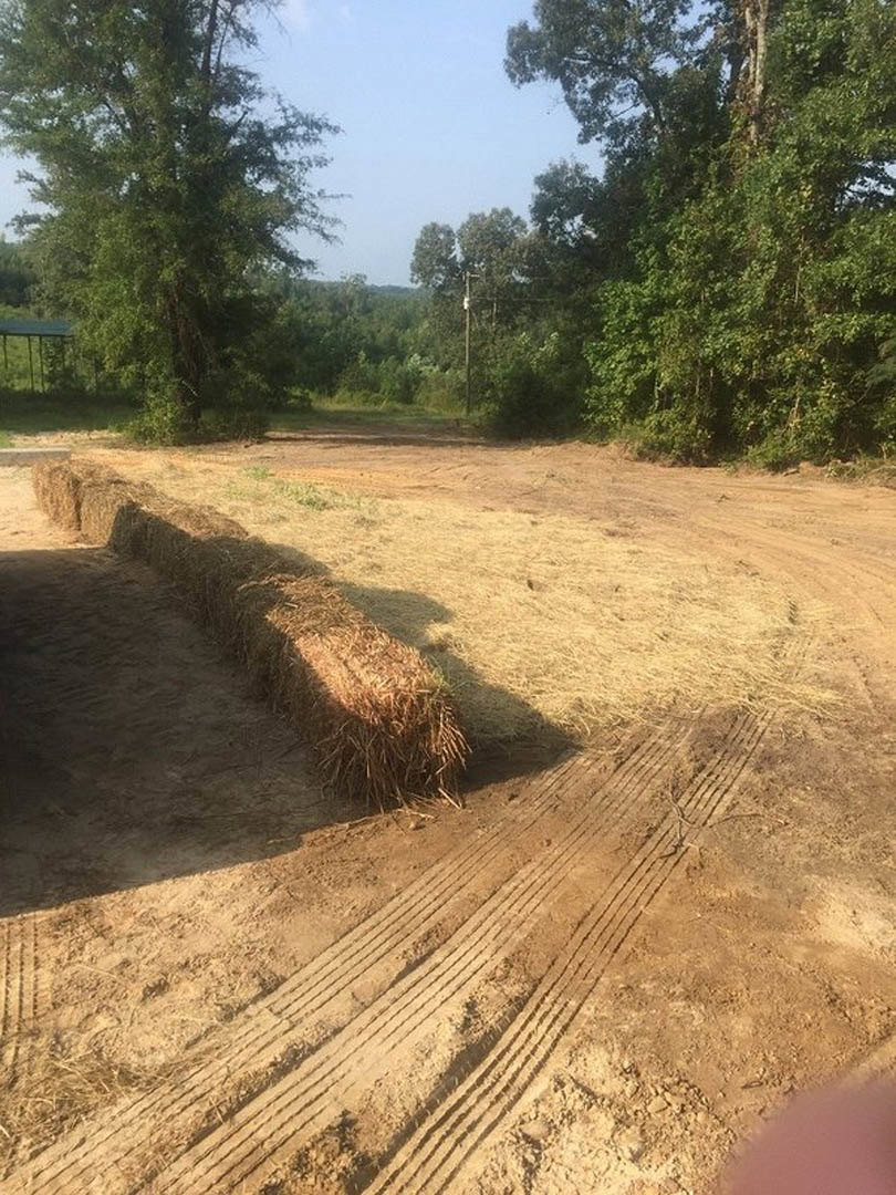 Expansive dirt field with scattered hay bale, bordered by leafy trees under clear blue sky