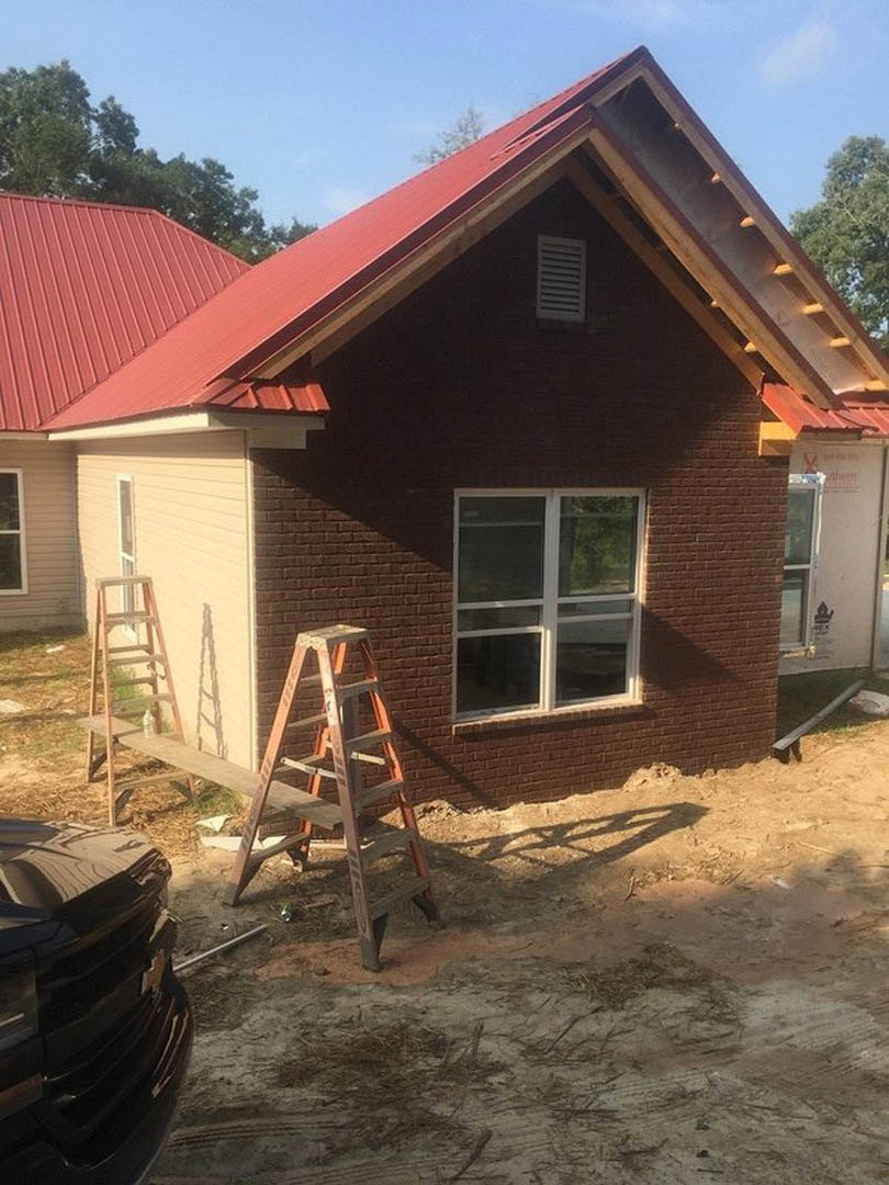 Red metal roof atop a brick house, white-framed windows with glass panes, exterior vent, ladder leaning against wall, two chairs on ground near front bumper of parked car, blue sky