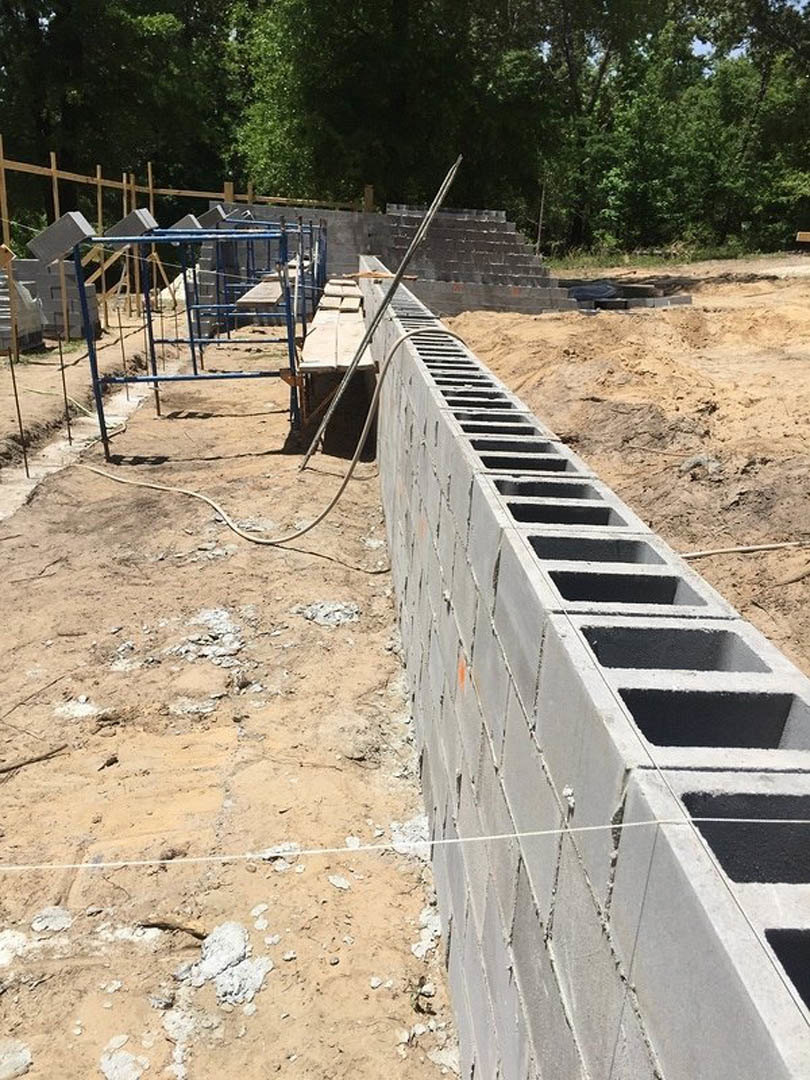 Concrete block wall under construction with visible holes, blue metal framing and poles, dirt ground marked with a white line, close-up of a white object, black rectangular item