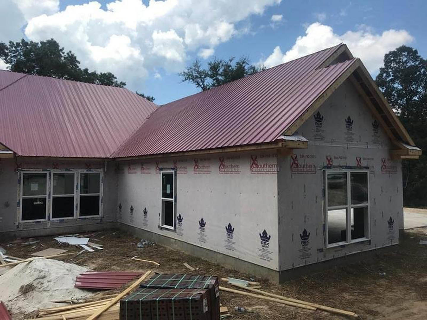 Partially built house with red metal roof, white-framed window, exposed brick walls, stack of bricks bound with rubber band, blue sky and clouds in background