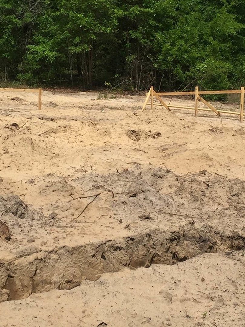 Dirt field bordered by a wooden fence, with mature trees in the background and scattered rocks on the ground