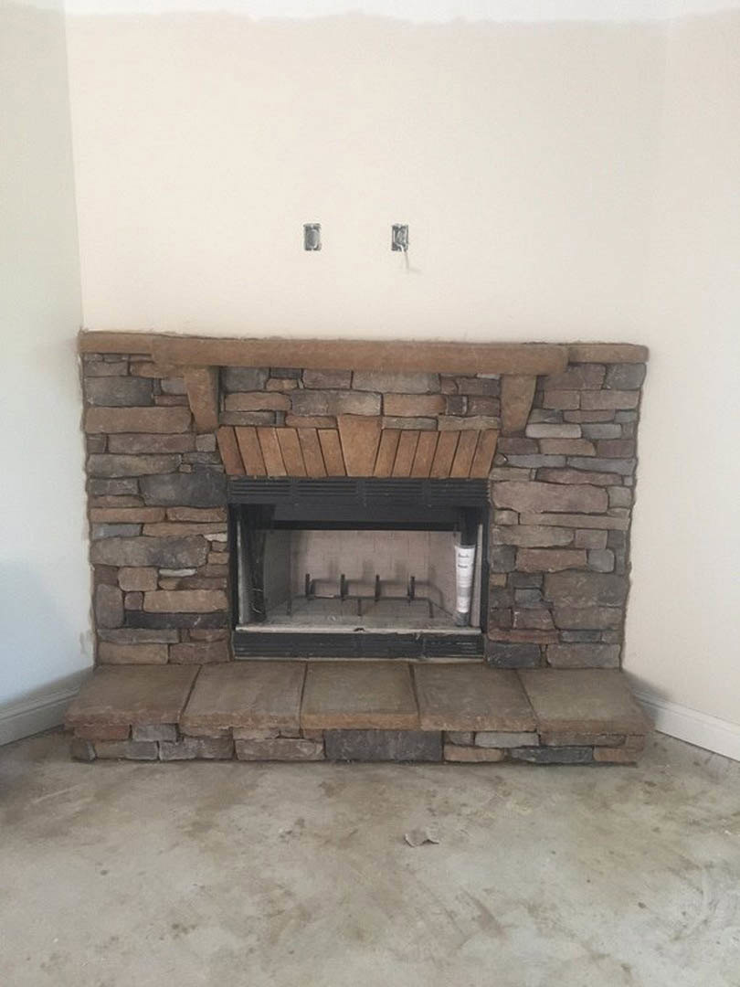 Stone fireplace with metal rack, square stone hearth, brick wall, concrete floor, and wood panel accents in a modern living room.