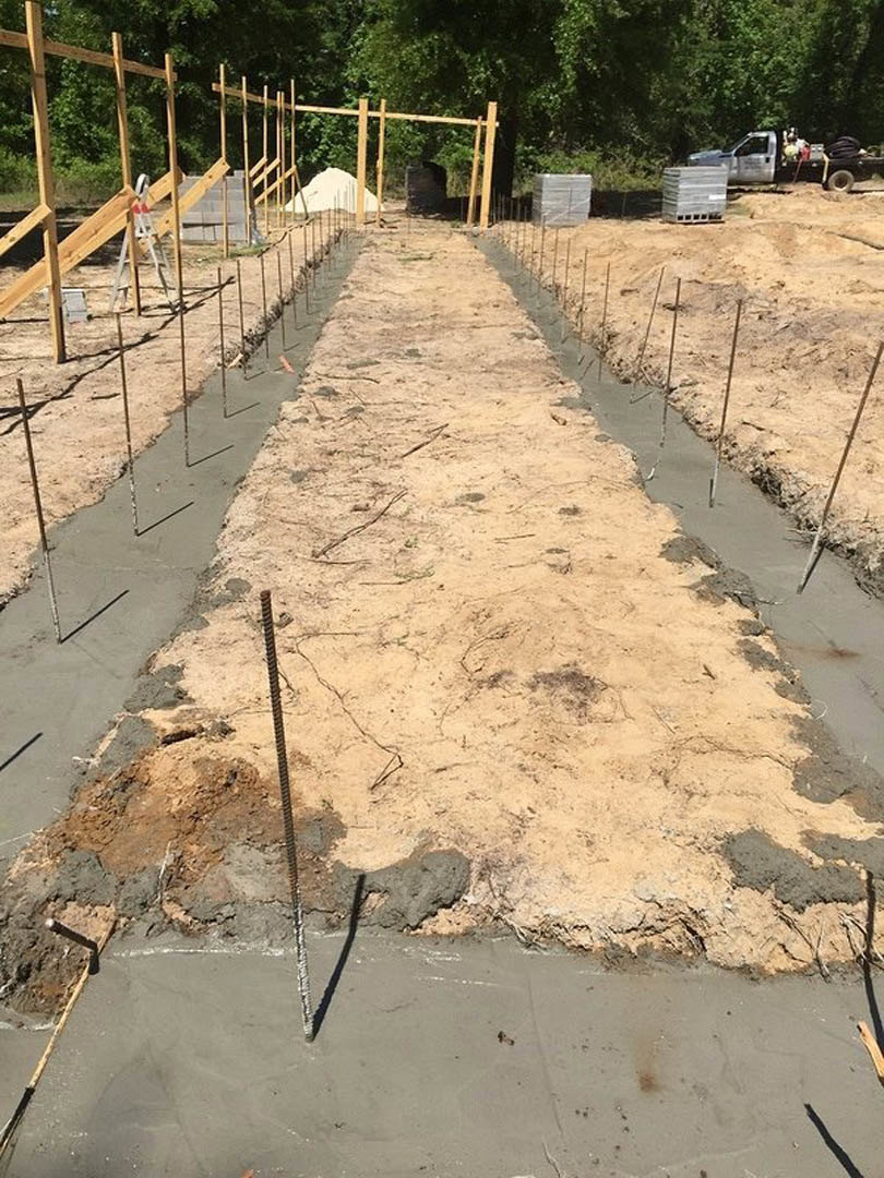 Concrete foundation under construction with metal rods protruding from the ground, wooden beams with ladders, stack of pallets, and a truck with people in the back; surrounded by