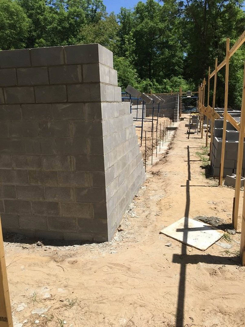 Concrete blocks stacked on a construction site, brick wall and metal fence in background, brick chimney partially visible with trees behind.
