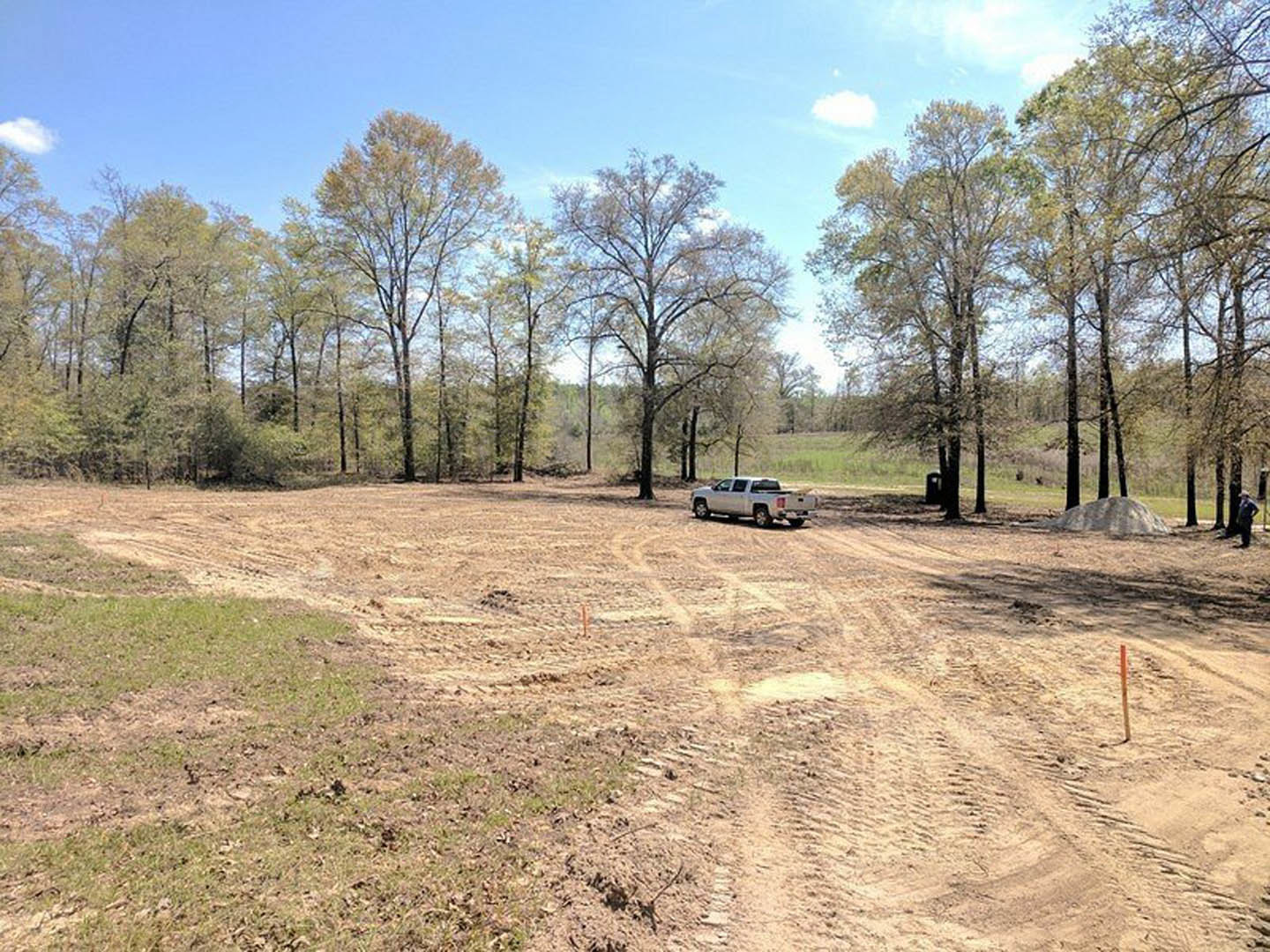 Silver pickup truck parked on a dirt field with scattered grass, bordered by trees under a partly cloudy sky