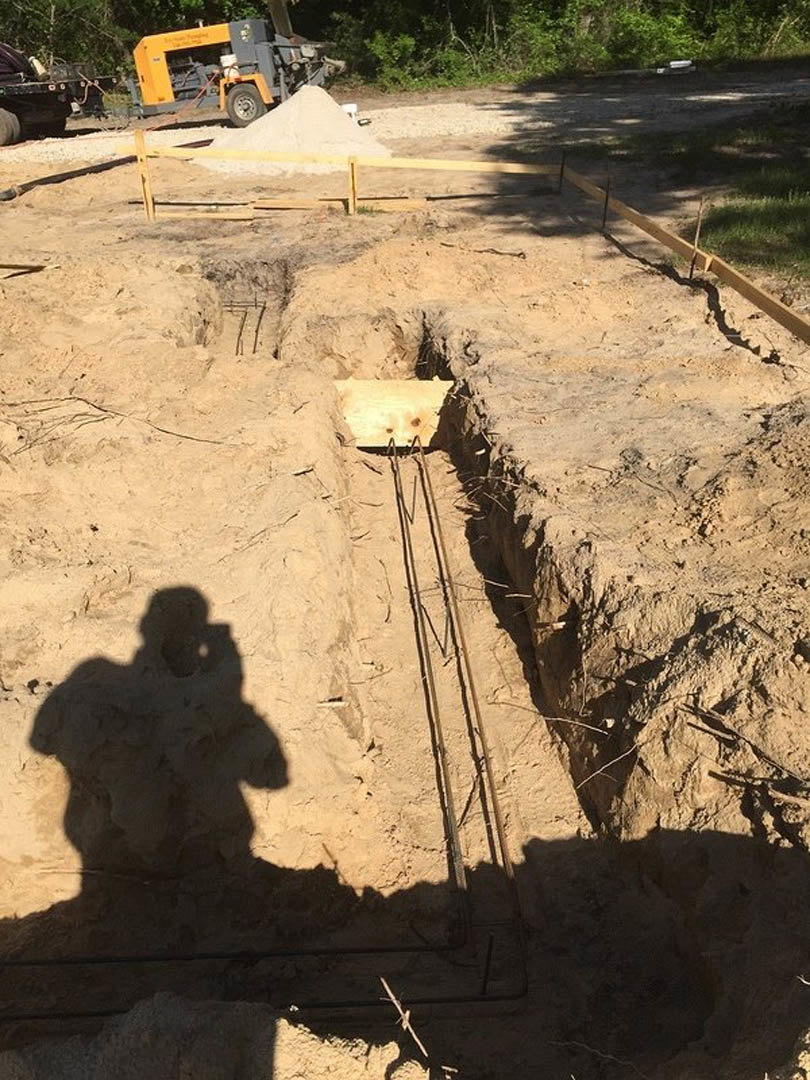 Freshly dug trench in sandy soil outdoors, yellow and grey construction equipment nearby, tree and plant visible in background, person’s shadow cast across ground.