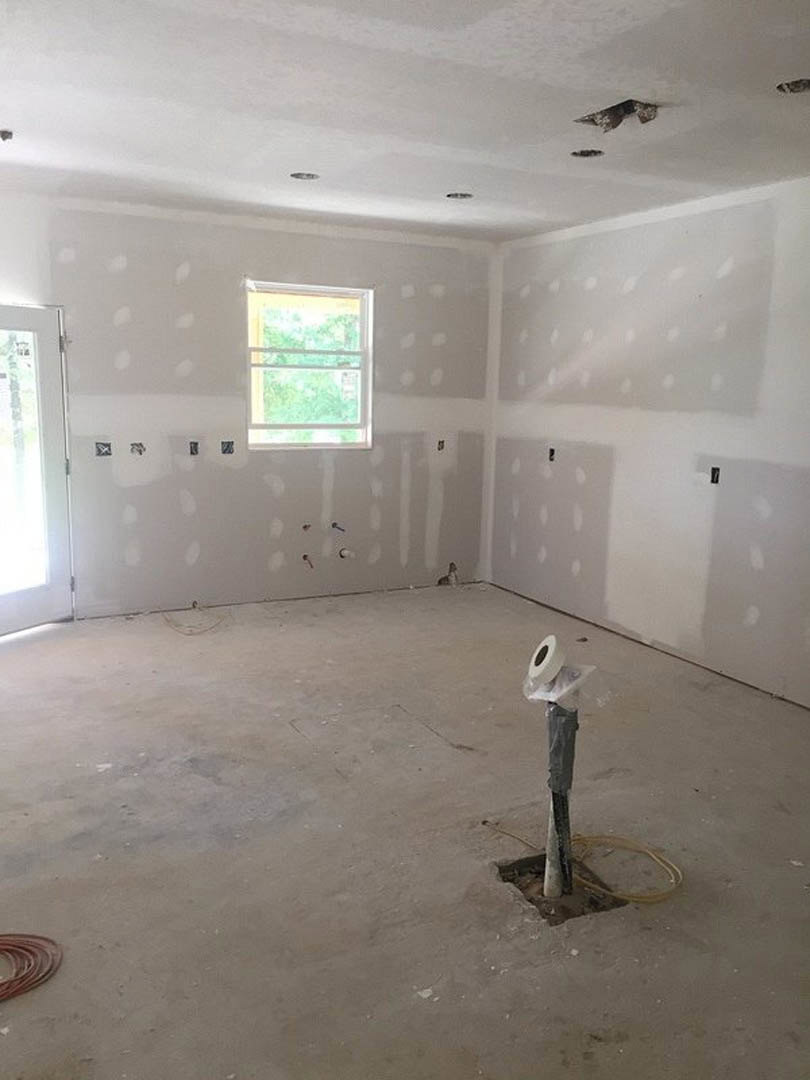 Unfinished room with plaster walls, exposed wiring, concrete floor, and a hole in the wall revealing pipes; white-framed window and white door with glass panel visible.