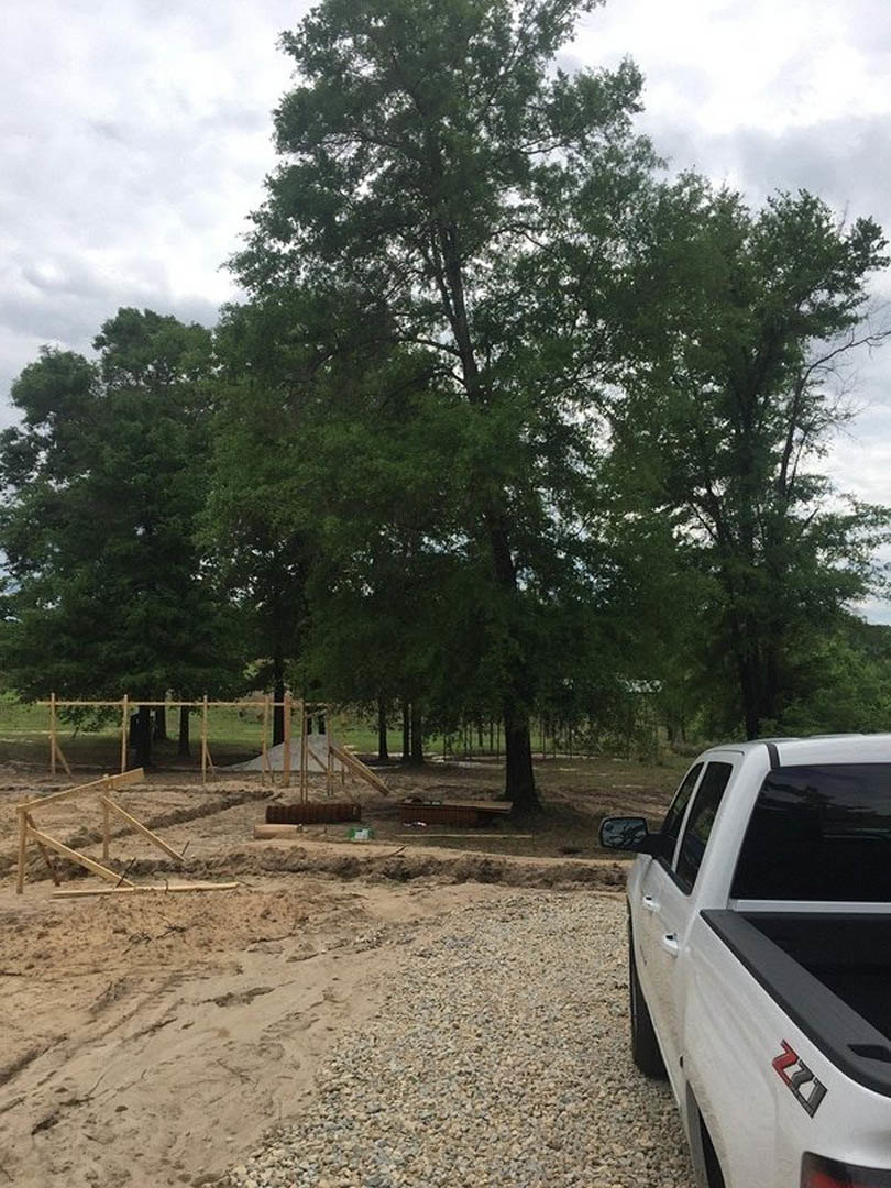 White pickup truck with black trim parked on gravel in front of partially built wooden-framed house, surrounded by dirt field and scattered trees under open sky