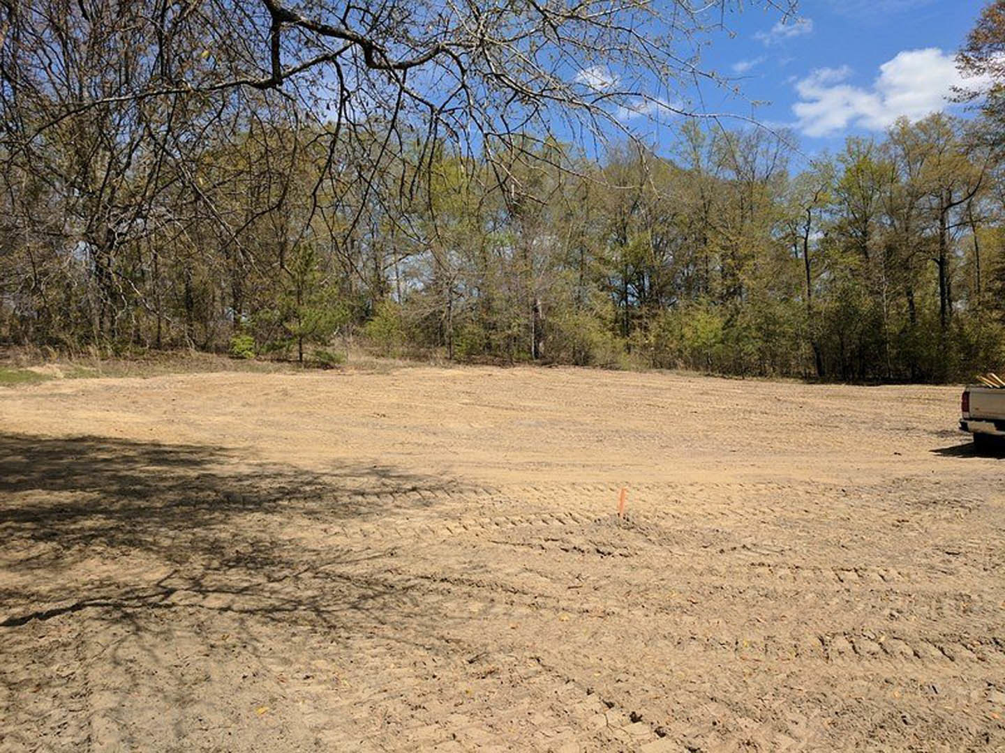 Dirt field with tire tracks, scattered trees, blue sky with clouds, partial view of white truck at edge of scene