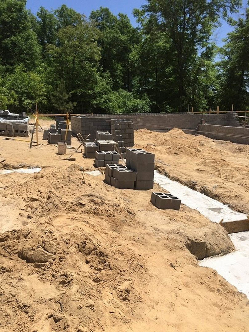 Cement blocks stacked on a dirt construction site, surrounded by piles of sand and grey bricks, with trees and a muddy road in the background