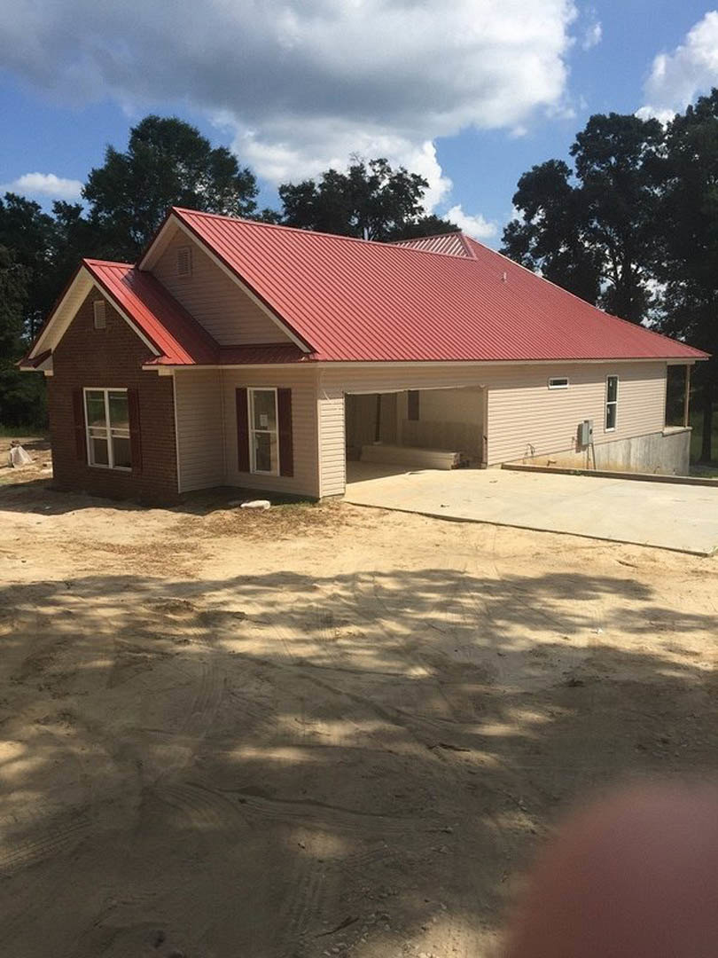 Red metal roof with triangular gables, white exterior walls, white-framed window, concrete slab foundation, dirt road with tire tracks, surrounding trees under cloudy sky.