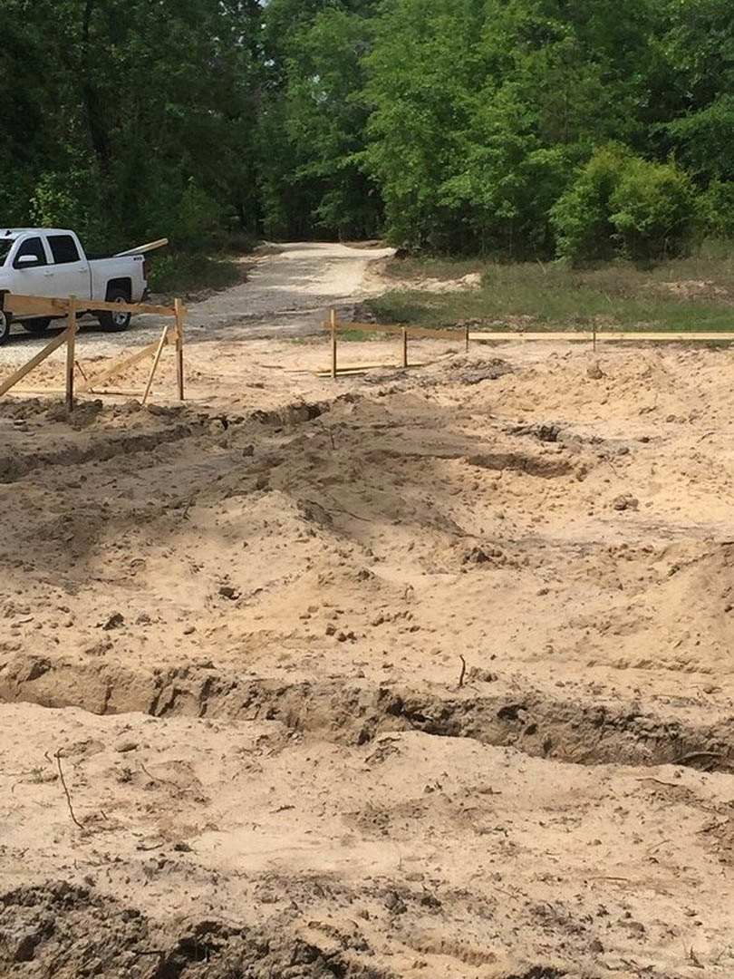 White pickup truck parked on a dirt lot with tire tracks, wooden fence along sandy edge, utility pole nearby, and dense trees in the background.