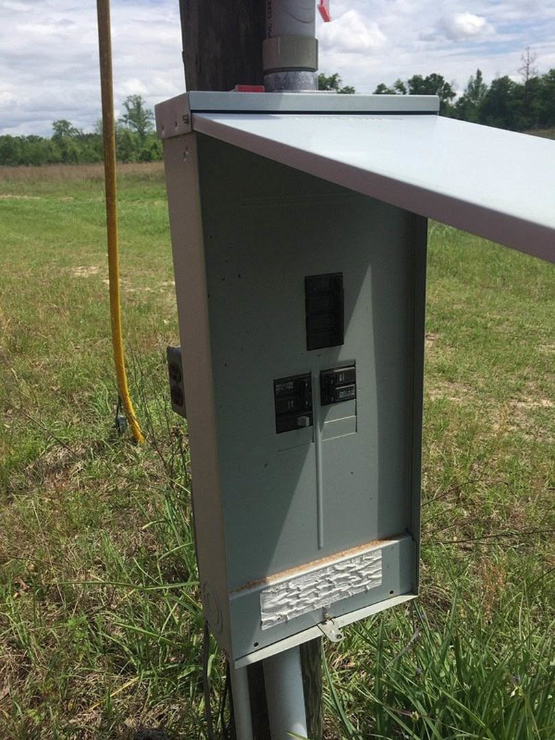 Black metal electrical box with multiple switches mounted outdoors near grass and trees under a cloudy sky