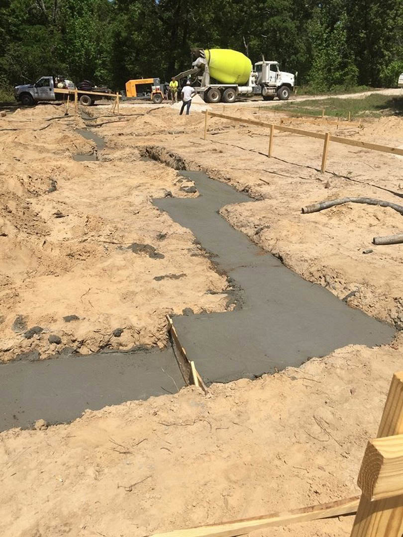 Cement mixer and construction materials on sandy ground, wooden fence, truck, and worker at custom home build site