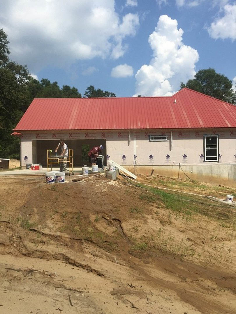 Man in red shirt painting white-framed window on house with red roof, buckets scattered on dirt hill, blue sky with clouds overhead.