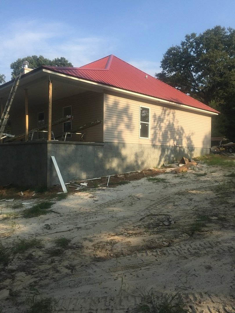 Partially built house with red metal roof, exposed framing, ladder leaning against unfinished wall, dirt road with tire tracks in foreground, tree nearby, cloudy sky overhead
