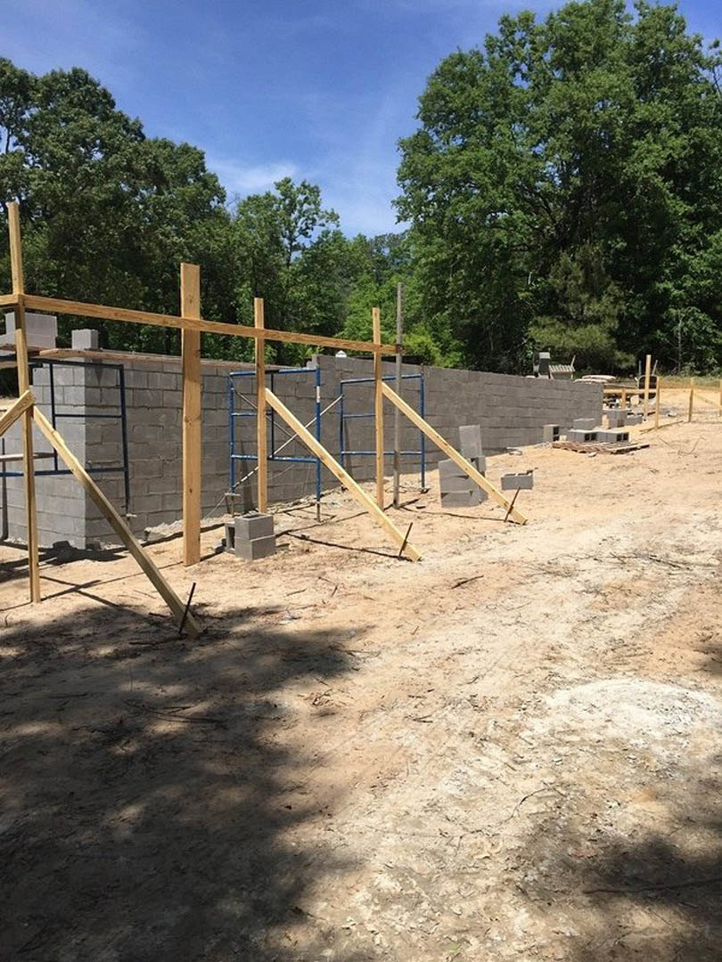 Partially built exterior wall with exposed brick and wooden beams, scaffolding set up alongside, large leafy tree and wood fence in background, cloudy sky overhead