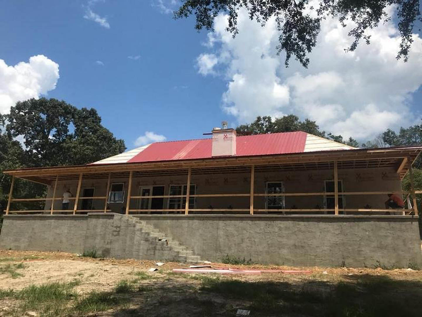 Red metal roof atop a white stucco house, concrete retaining wall with stairs, landscaped yard, partly cloudy sky.
