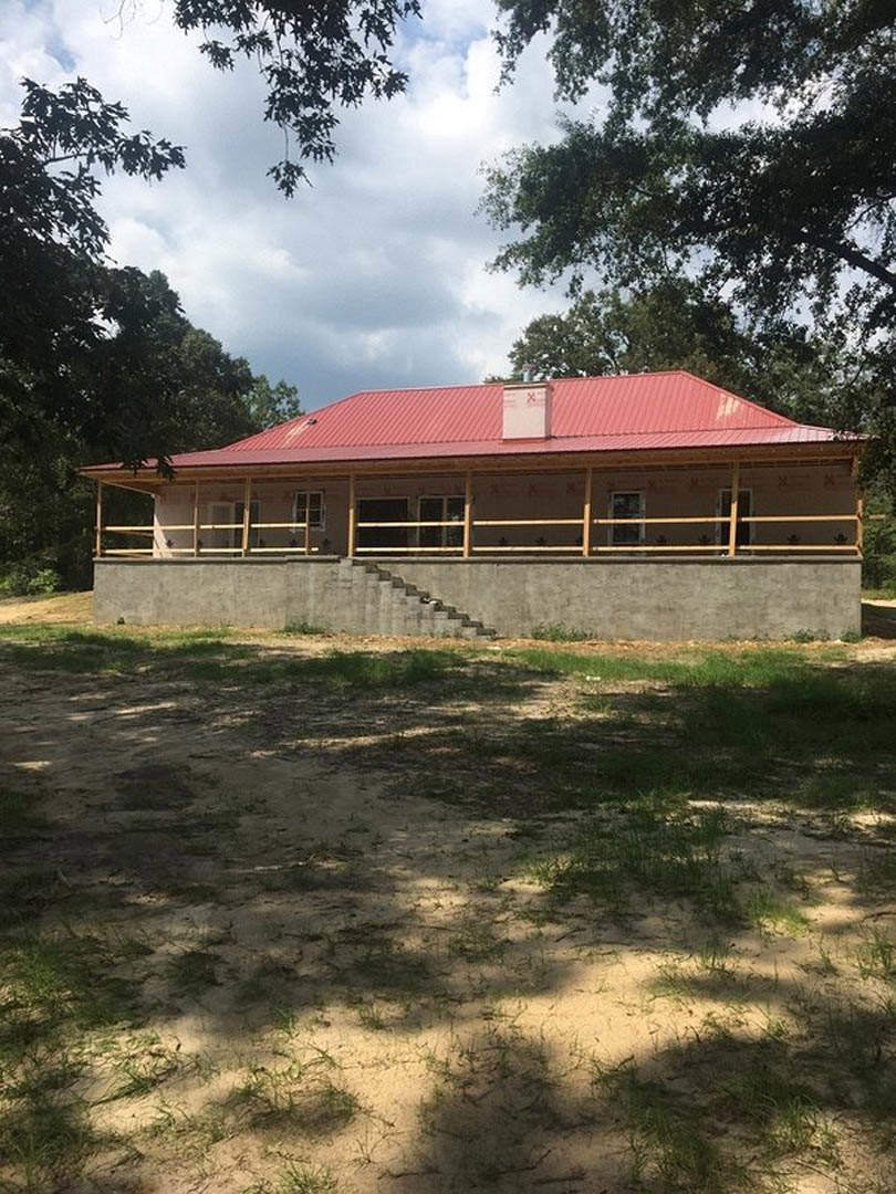 White stucco house with a red gabled roof and chimney, concrete steps leading to entry, grassy yard bordered by a stone retaining wall, large windows, mature trees, and partly