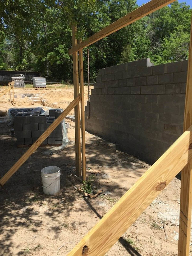 Partially built home with exposed wooden framing, stacked lumber on pallets, concrete block wall, bucket of bricks, and construction materials scattered on dirt ground