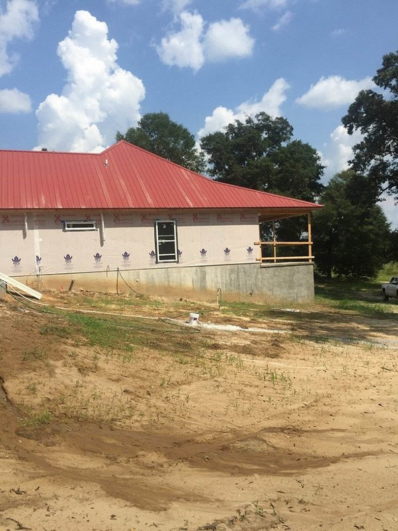 Red metal roof atop a white house with white-framed windows, concrete boundary wall, patchy grass and dirt yard, cloudy sky, and surrounding rural landscape.