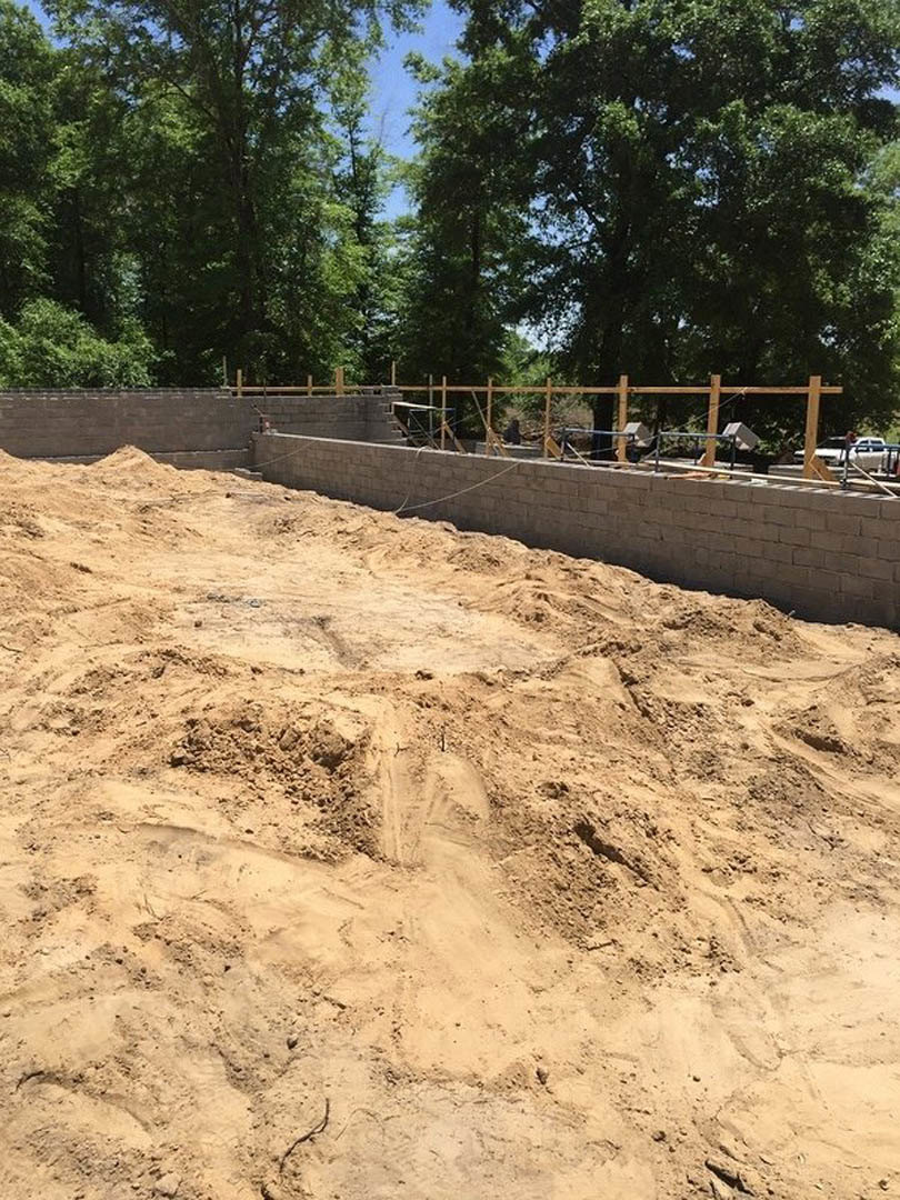 Dirt and concrete retaining wall bordered by trees, large sand pile in foreground, man balancing on rope, fence and utility box visible, person walking up stairs in background