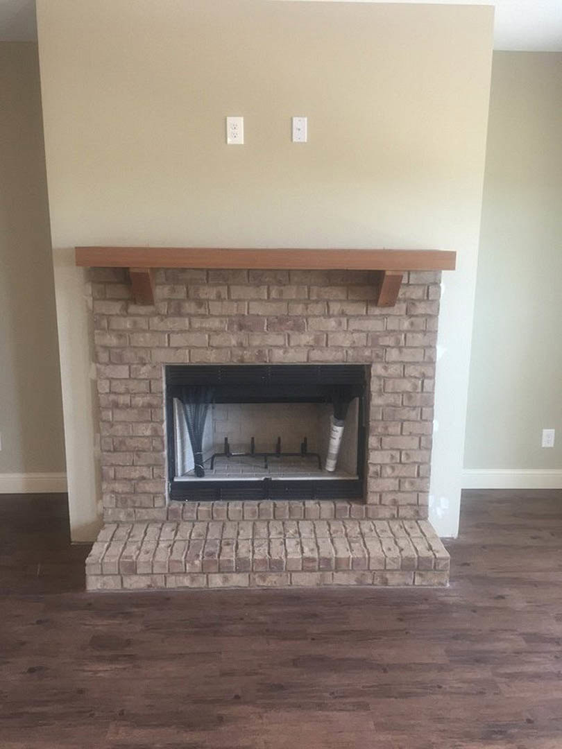 Black-framed fireplace with wood beam mantel set against a brick wall, wood flooring and brick step in foreground