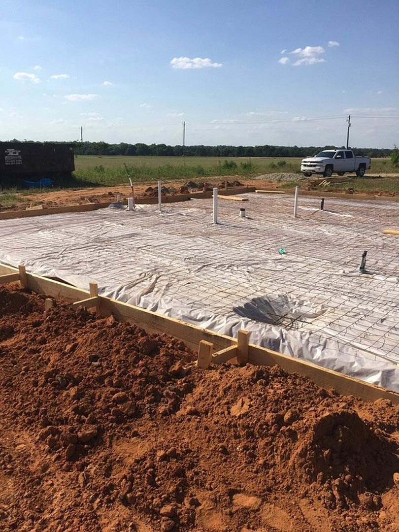 Foundation construction site with exposed soil, wood framing, plastic sheeting, metal fencing, white pickup truck parked roadside, dirt piles, and partly cloudy sky