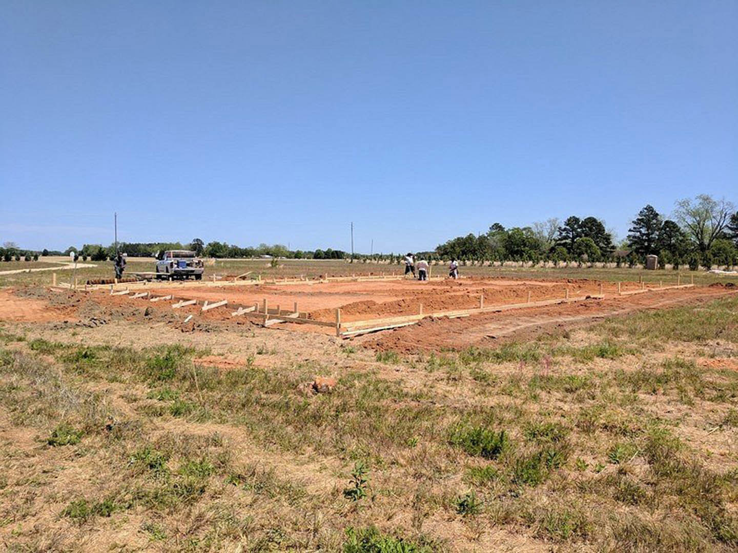 Workers preparing soil and grass on rural construction site, white truck parked roadside, fenced area in foreground, scattered trees under partly cloudy blue sky