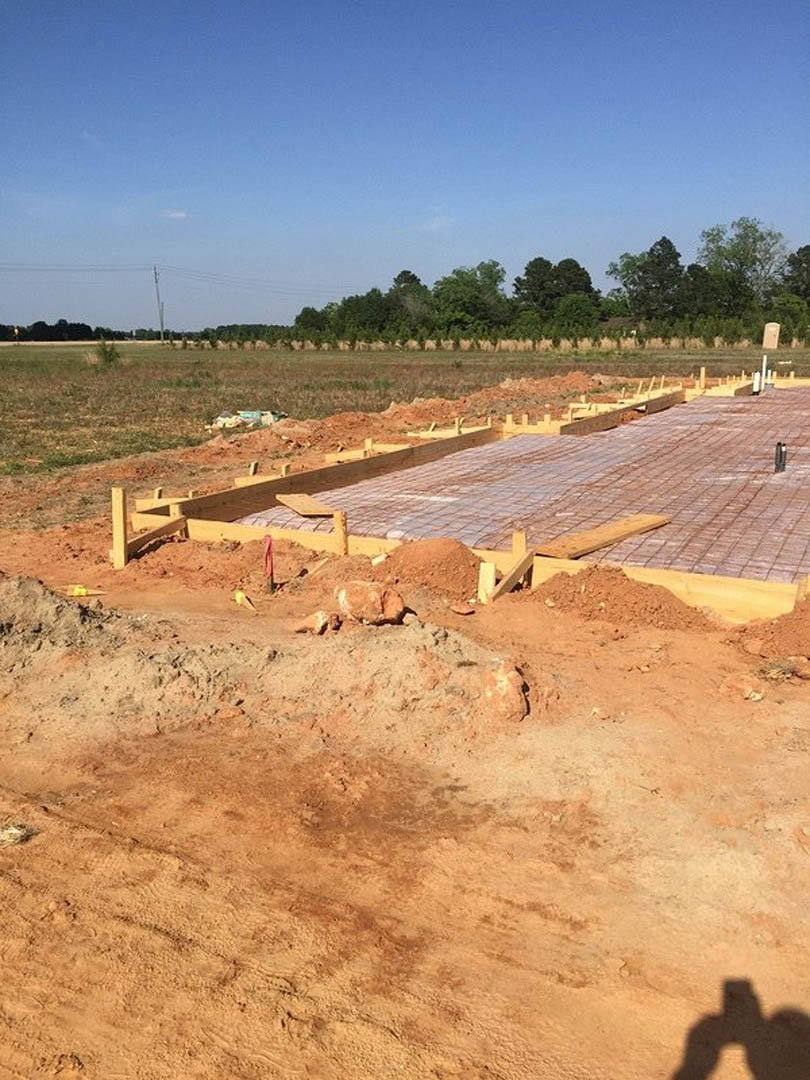 Concrete foundation with wooden framing at a residential construction site, dirt piles and wooden fence in foreground, group of trees and blue sky with clouds in background.