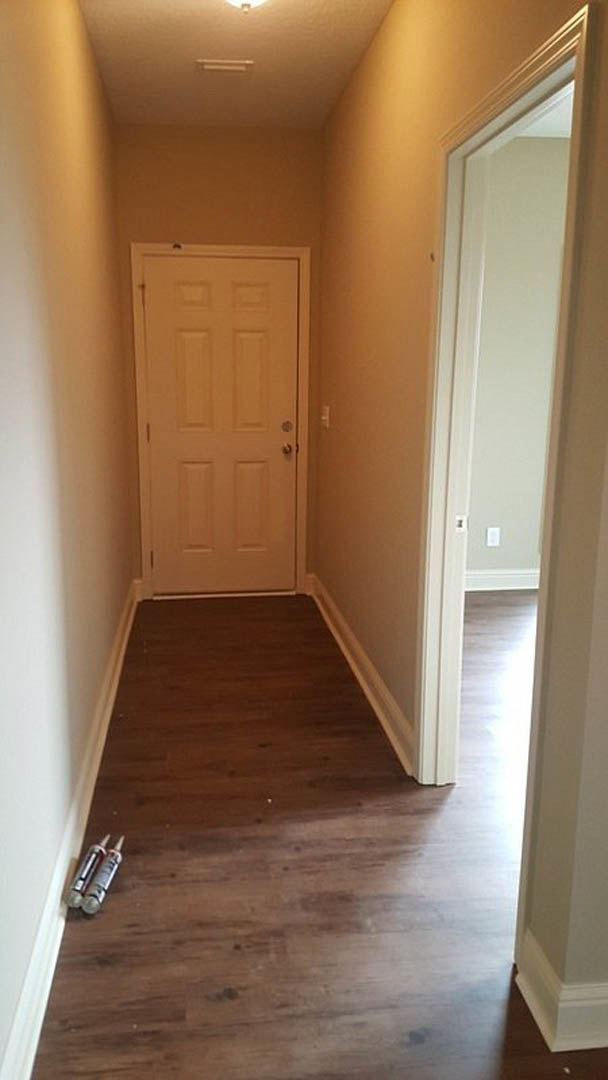 White hallway with hardwood floor, white door featuring gold handle, ceiling light fixture, two bottles and glue containers placed on the floor near the doorway