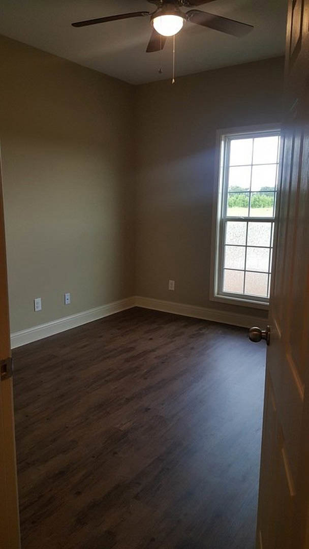 Bright room with dark wood flooring, white plaster walls, large window with grid framing and view of trees, ceiling fan with light fixture.