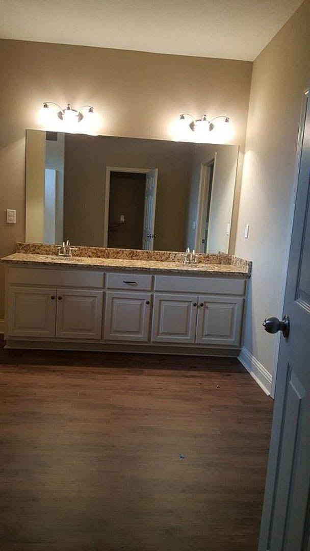 Bathroom with expansive wall mirror, wood flooring, marble countertop, white cabinetry, tile backsplash, and door with light switch visible.