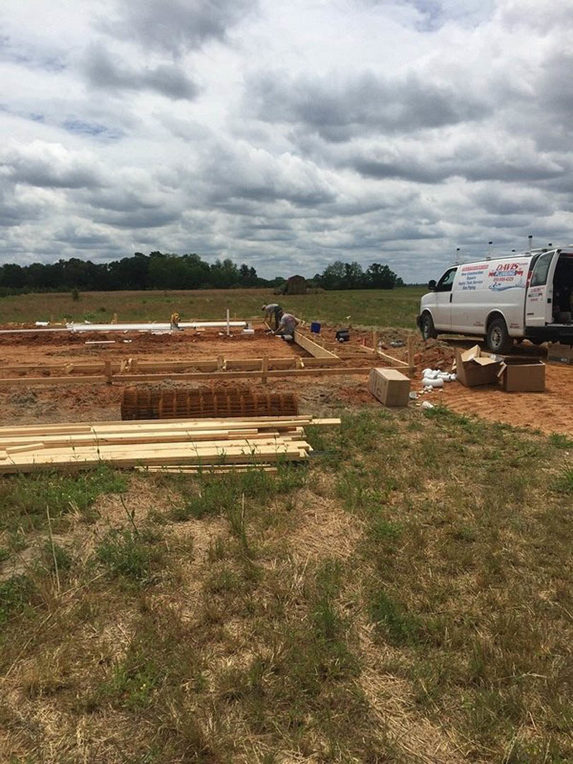 White van parked on grassy lot beside scattered lumber, cloudy sky overhead, trees in background, wooden step and cardboard box visible in foreground.
