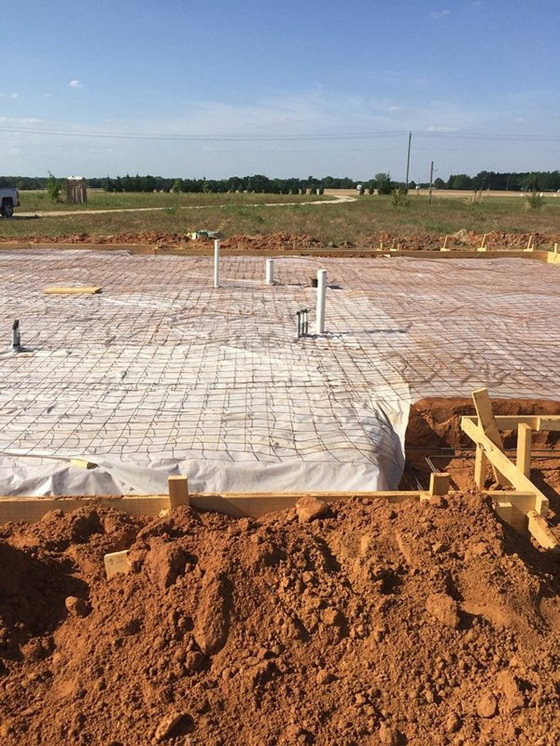 Exposed soil and metal rebar at a residential foundation construction site, wooden formwork and white poles under a partly cloudy blue sky