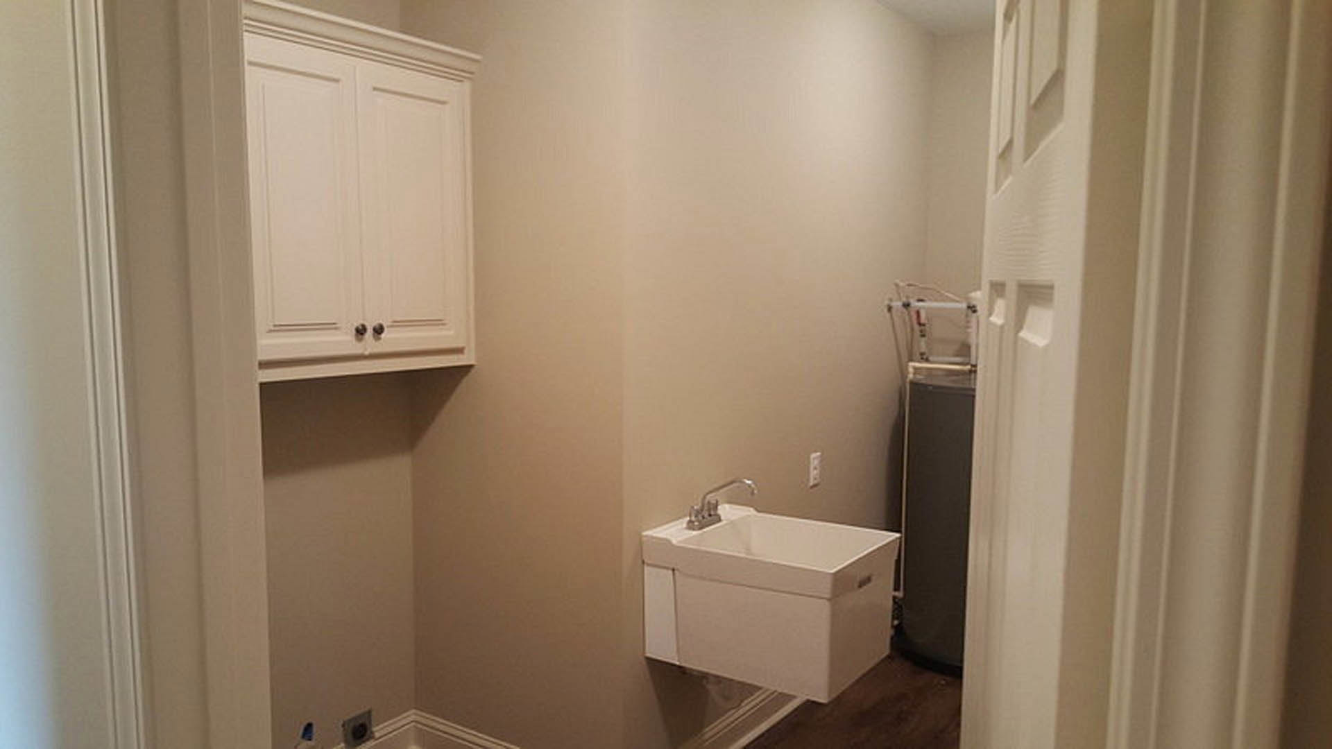 White laundry room sink with chrome faucet, set in a tiled countertop beneath white cabinets and surrounded by light-colored walls.