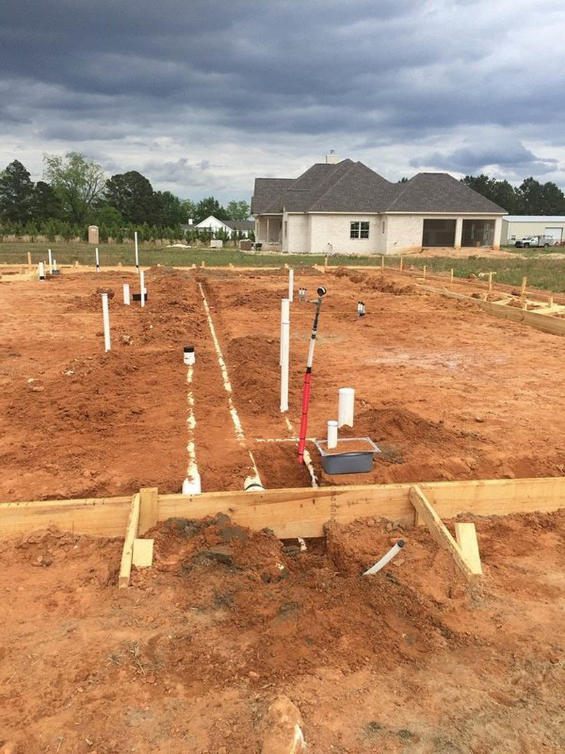 Partially built house with exposed wooden framing and black roof, construction materials scattered on dirt lot, completed home and cloudy sky in background