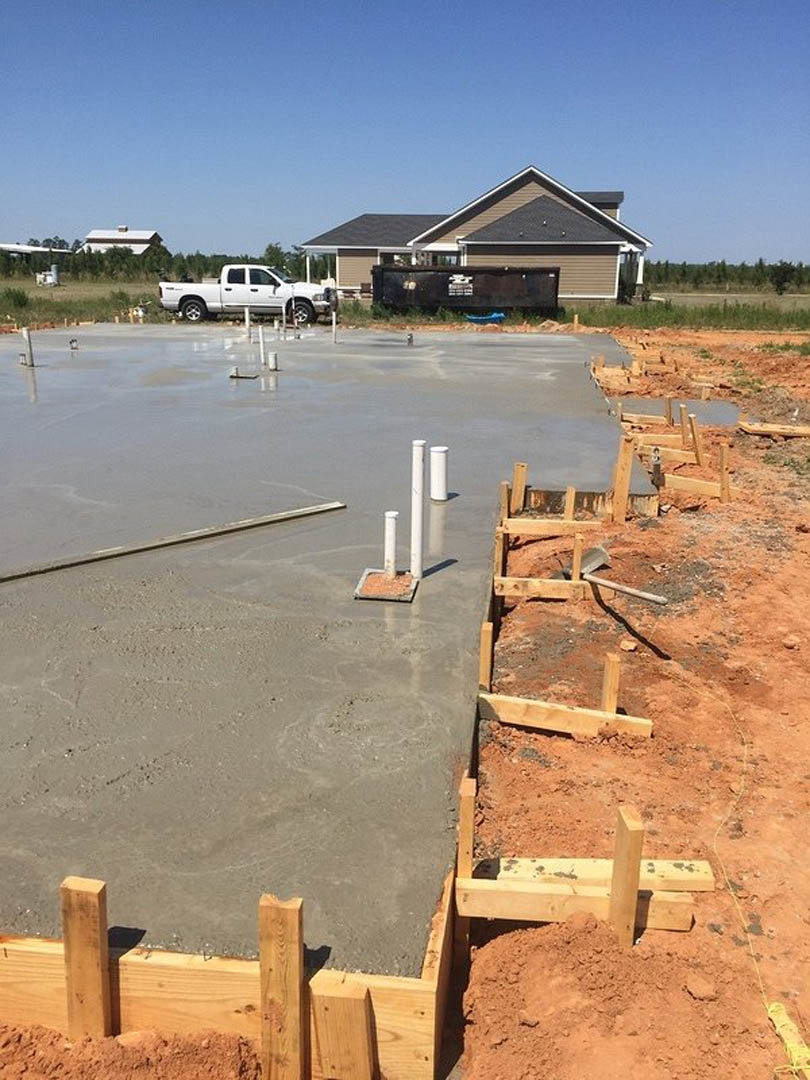 Partially built house with exposed wooden framing, construction materials scattered on dirt ground, white truck parked nearby, wooden fence with central opening, metal pole lying