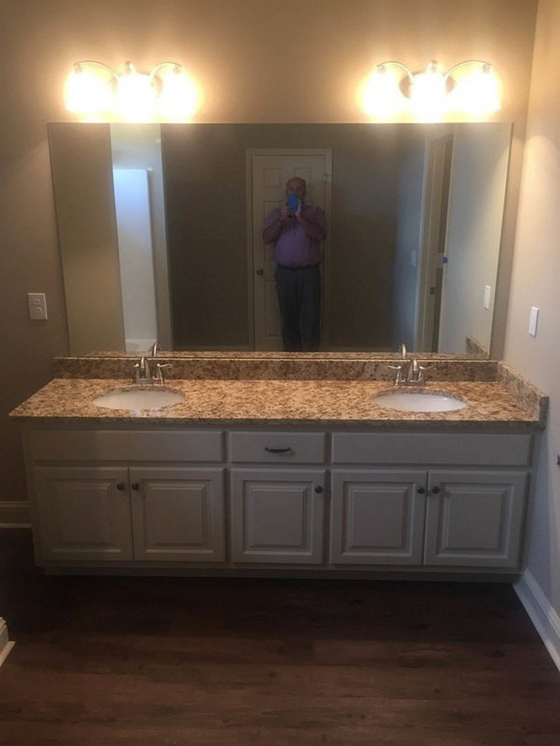 Man in purple shirt taking a selfie in bathroom mirror, stone countertop, wood cabinetry, tile walls, chrome faucet, overhead lighting