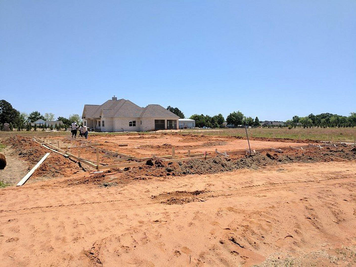Large residential foundation surrounded by dirt, partially constructed house with roof, workers in safety vests, blue sky overhead, trees in background