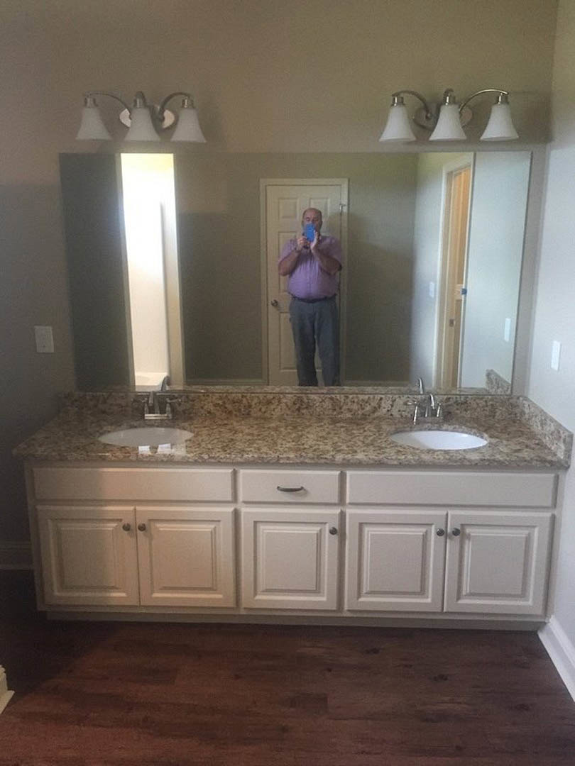 Man taking a selfie in bathroom with wood floor and white border, stone countertop, white cabinetry, and chrome plumbing fixtures