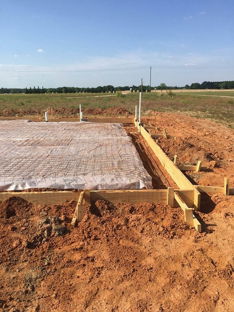 Wood and metal foundation under construction with exposed beams, dirt and wood framing, tarp covering materials, distant trees, and partly cloudy blue sky