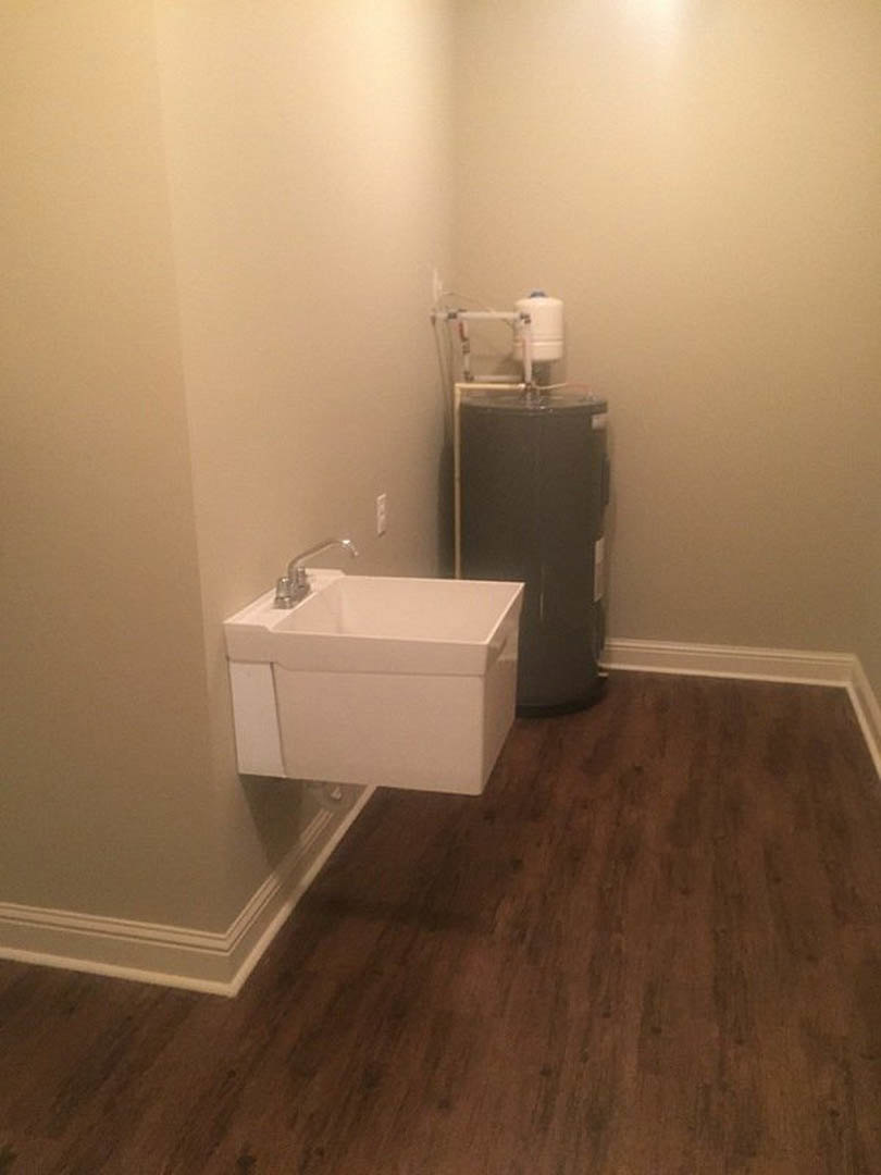 White ceramic sink with chrome faucet beside a tankless water heater mounted on plaster wall, wood flooring, and tile backsplash in utility room.