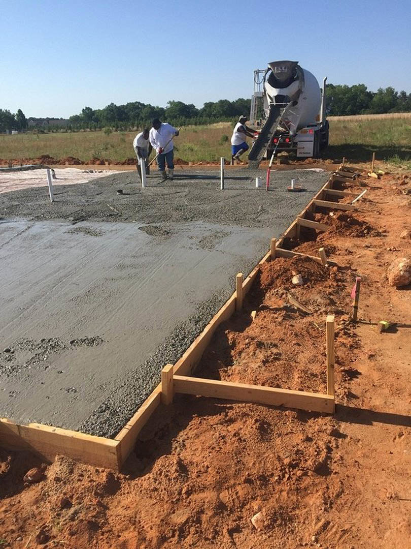 Workers spreading and leveling wet concrete on a foundation slab at a residential construction site, cement mixer truck nearby, blue sky overhead, trees in background
