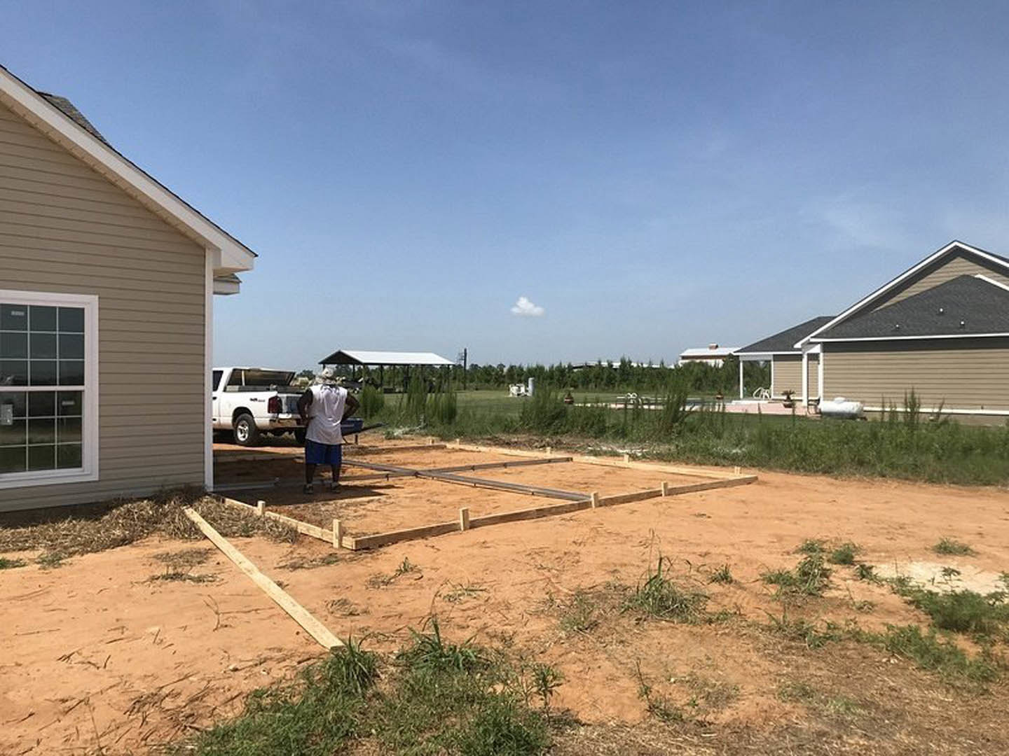 Man in white shirt standing in front of modern home with white-framed windows, wooden fence, white pickup truck with open trunk, dirt yard, and cloudy sky