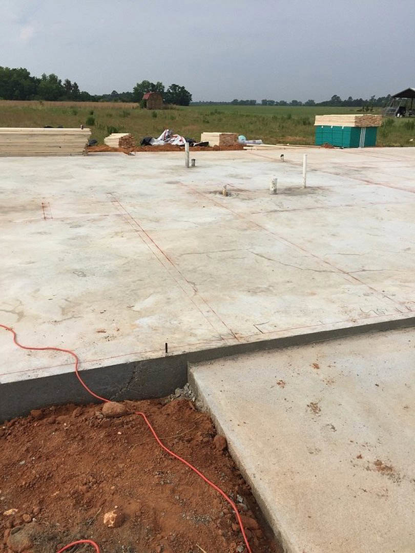 Concrete slab with red rope lying across the surface, surrounded by red dirt, green and white wall in background, trees and building visible outdoors under cloudy sky.