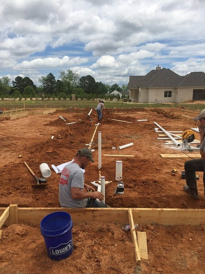 Workers installing pipes and building foundation near a partially constructed house with chimney, surrounded by soil, trees, and scattered construction materials under a cloudy sky